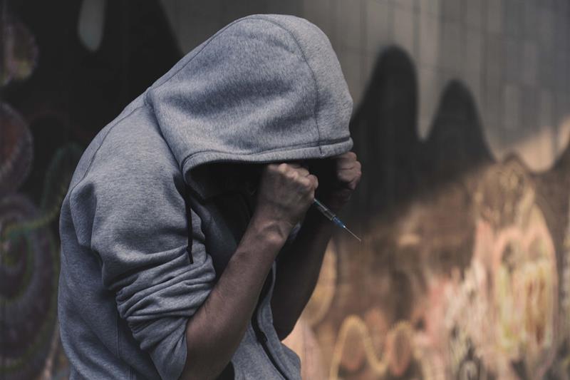 Person in a gray hoodie with hood up, head bowed and hands holding a syringe against their arm beside a graffiti-covered wall. Anxiety vs Anxiety Disorder